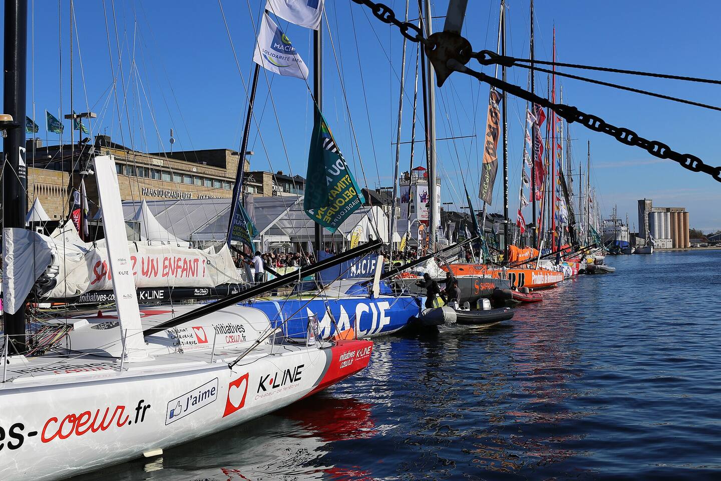 En flotta segelbåtar av IMOCA-klass förtöjda i en hamnbassäng före start